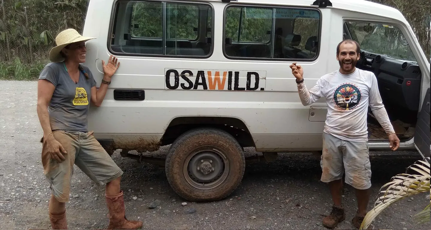 Two muddy people standing and smiling beside a white van with the word OSWILD on the door.