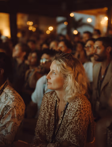 Focused woman with blonde hair and leopard print top sitting among an audience in a dimly lit setting.