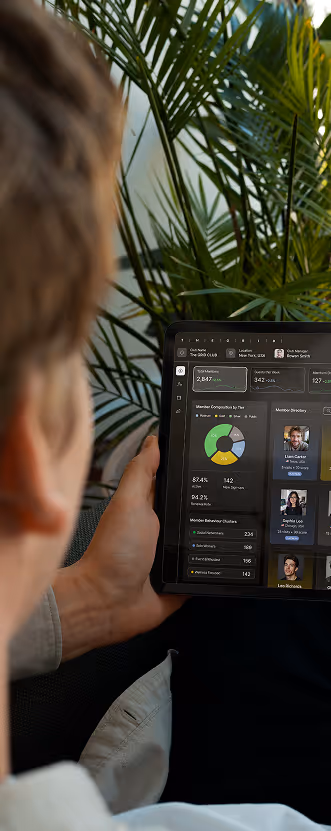 Person holding a tablet displaying a dashboard with graphs, member directory photos, and operational statistics set against a backdrop of green palm leaves.