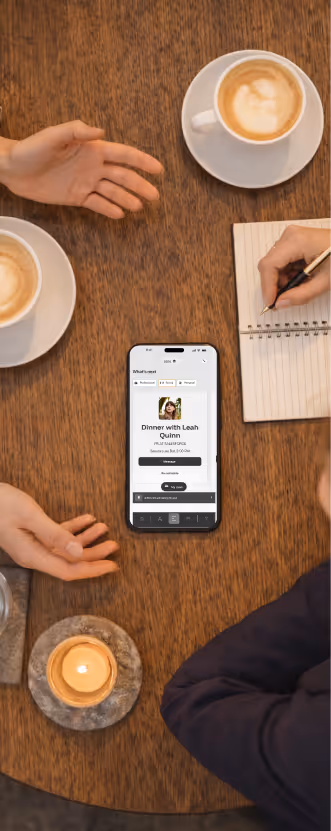 Top view of a wooden table with two cups of coffee, a lit candle, a smartphone displaying a dinner invitation, and two people with one taking notes on a notepad.
