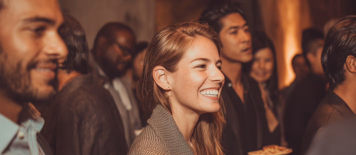 Smiling woman in a brown coat at a lively social gathering with diverse people in the background.
