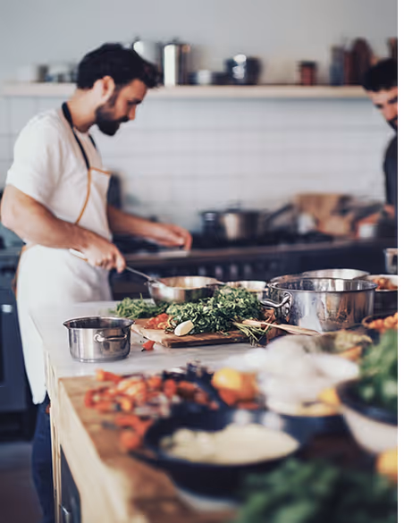Two men cooking in a kitchen, with fresh herbs and vegetables on a counter in the foreground.