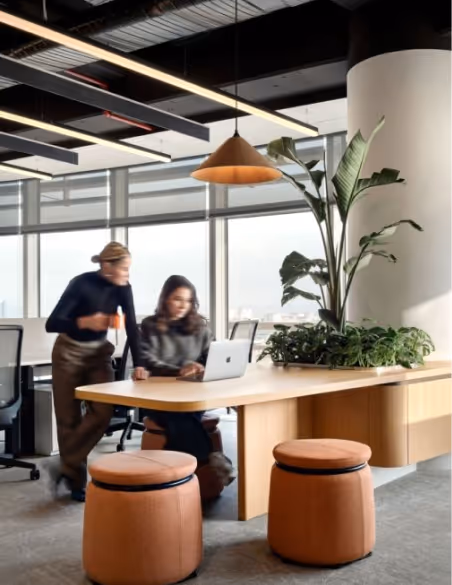 Modern office space with two women working together at a wooden desk with a laptop, surrounded by plants and orange cushioned stools.