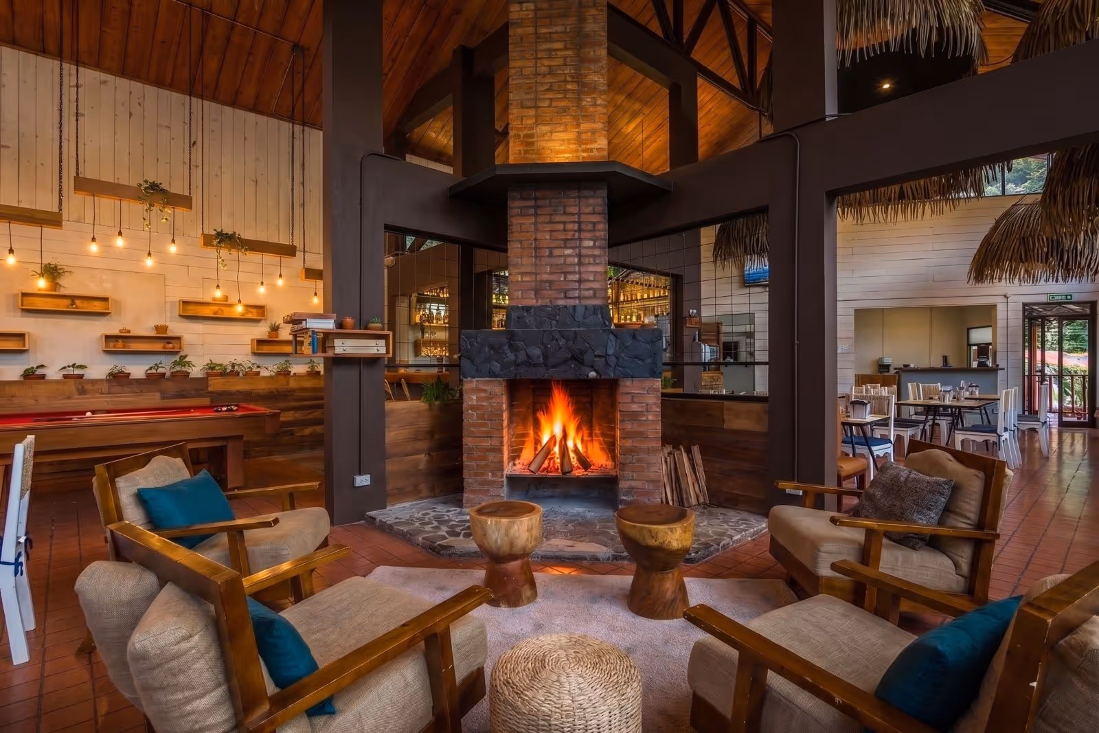 Cozy indoor lounge area with wooden armchairs and cushions around a lit brick fireplace in a rustic hotel interior.