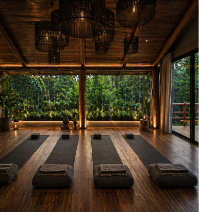 A serene yoga retreat space with four black mats and cushions on a wooden floor, surrounded by large windows showcasing lush greenery outside.