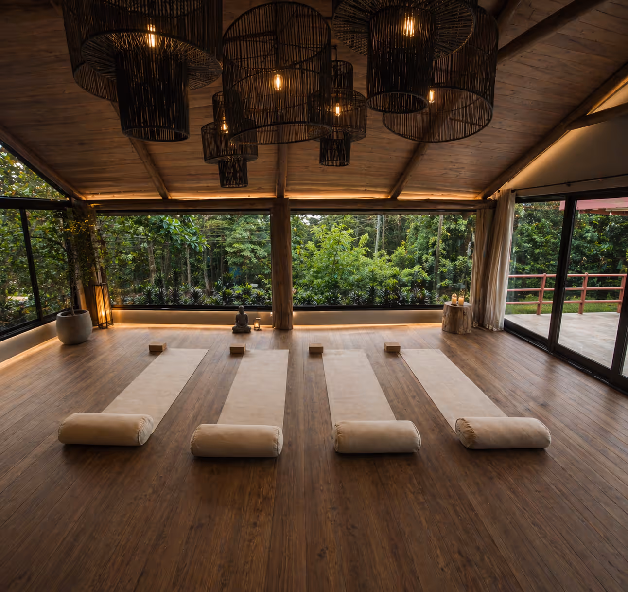 Yoga studio with wooden floors, four beige yoga mats with bolsters and blocks, large windows showing green trees outside, and hanging black lanterns.