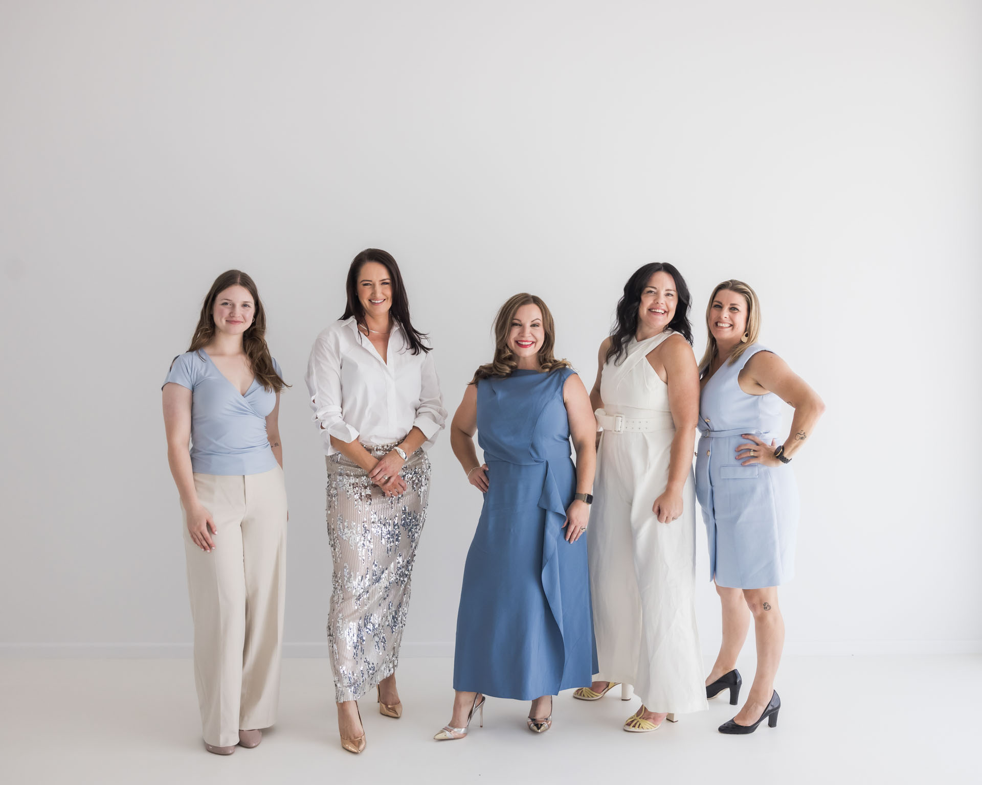 Five women in stylish outfits standing against a white background, smiling at the camera.