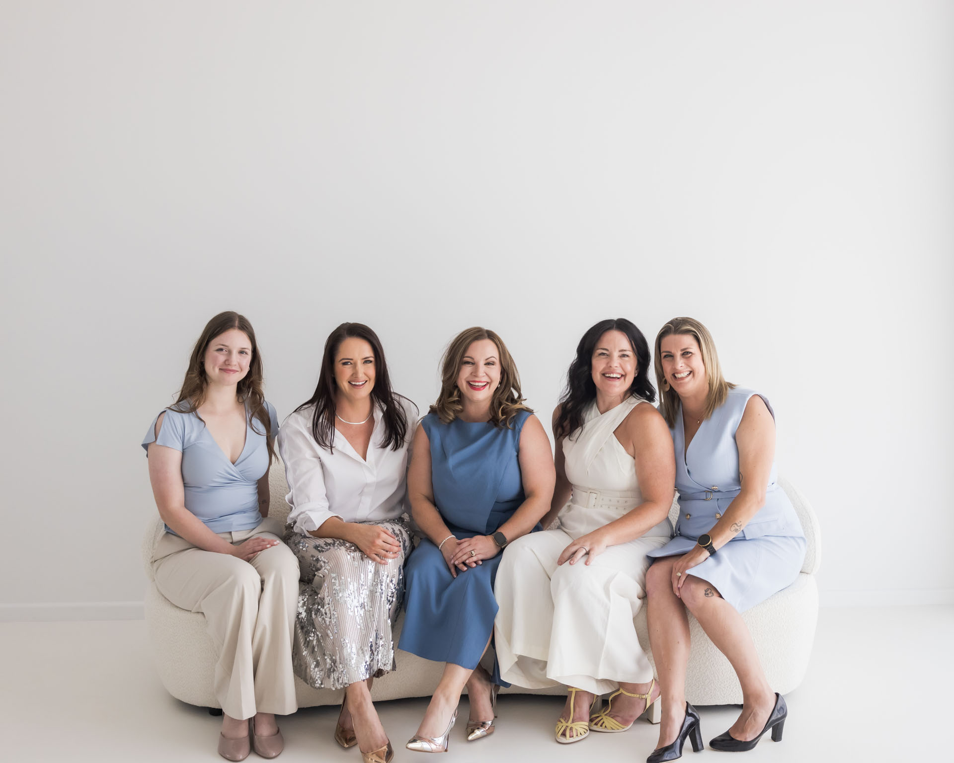 Five women sitting closely on a white couch against a plain white wall, smiling at the camera.
