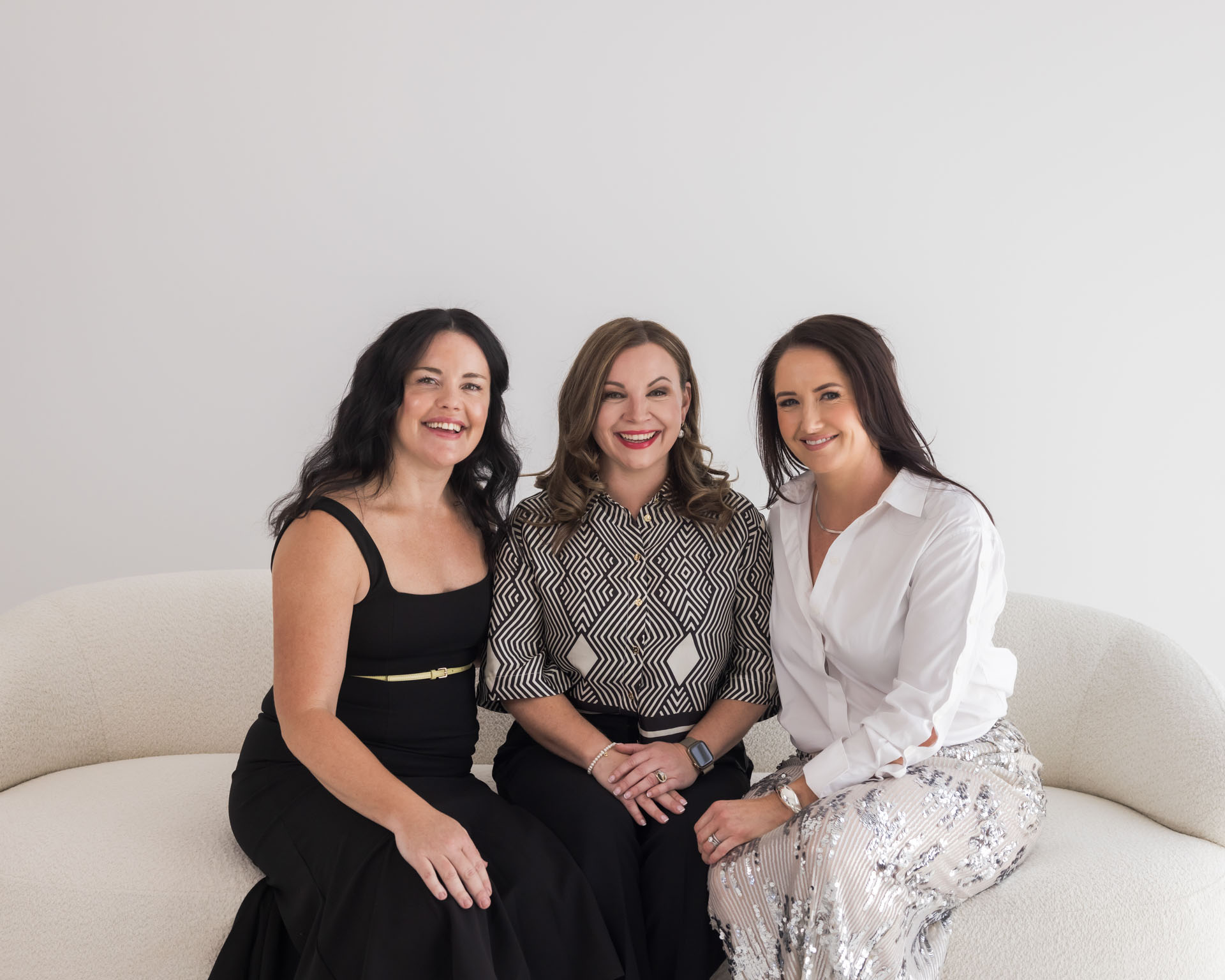 Three women sitting closely on a white sofa, smiling at the camera against a plain white background.