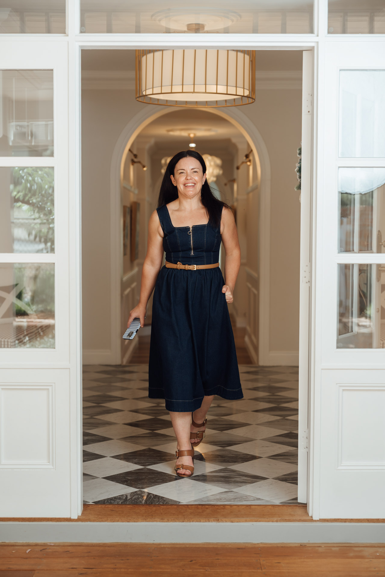 Smiling woman in a sleeveless navy dress and brown sandals walking through doorway indoors.