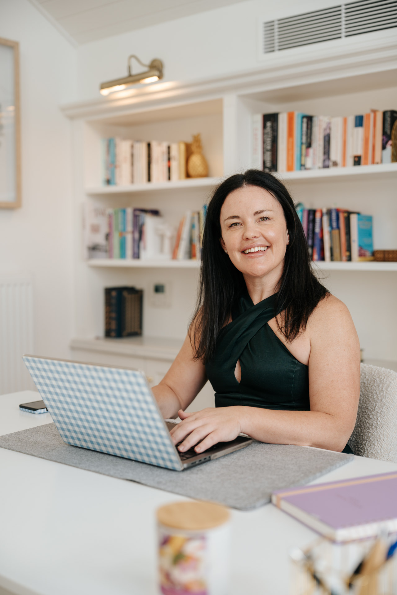 Smiling woman in black sleeveless top working on a laptop with a blue checkered cover at a white desk.