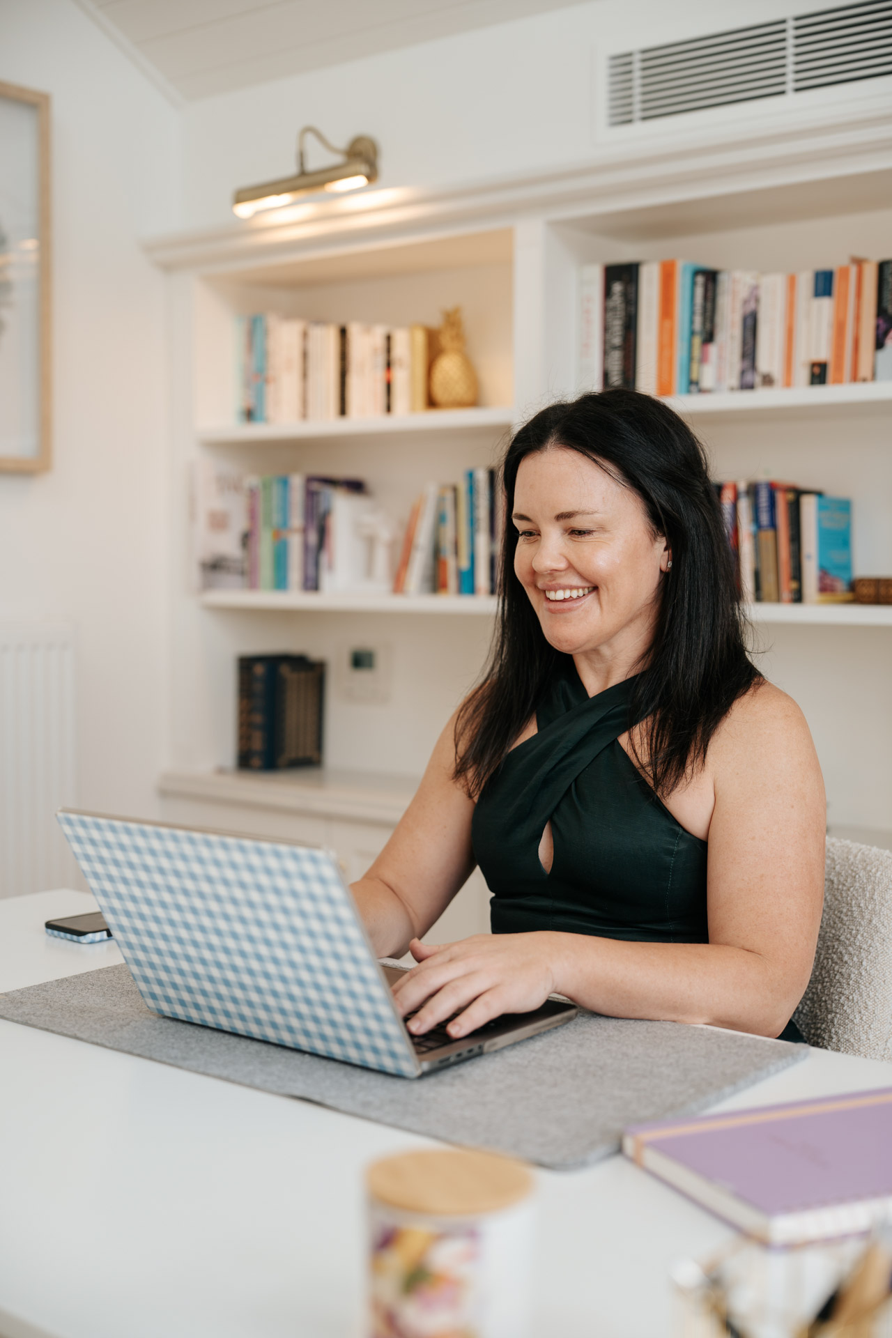 Smiling woman with black hair working on a laptop at a desk with bookshelves behind her.