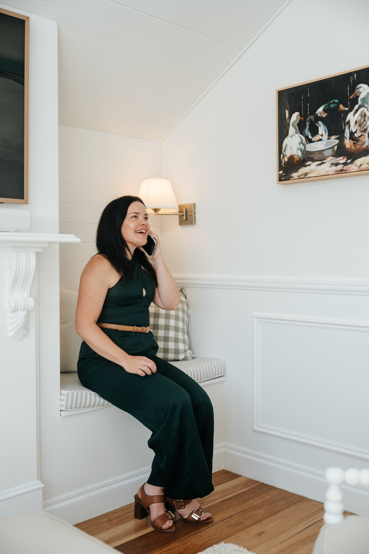 Woman in black dress sitting on a built-in bench talking on a smartphone in a white room.