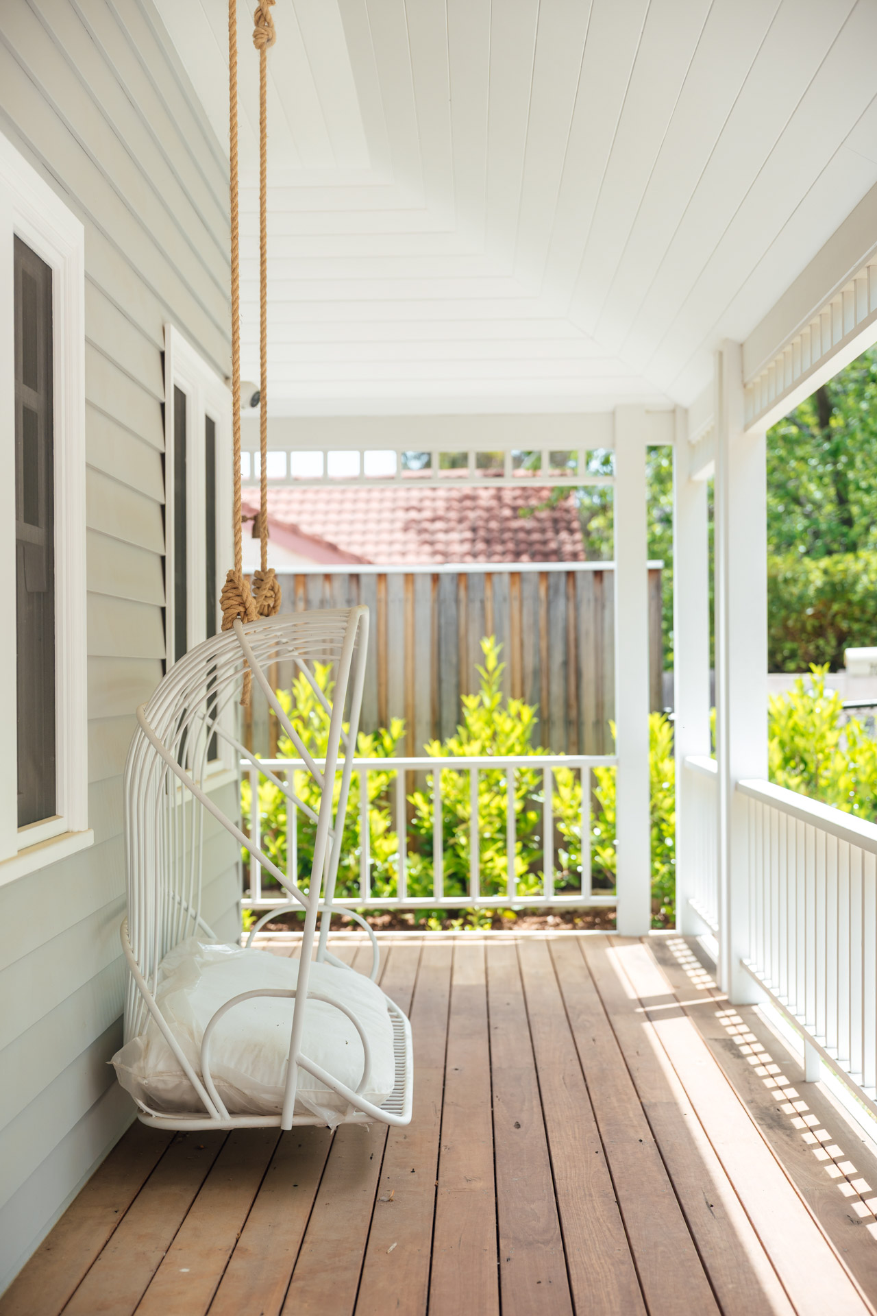 White hanging swing chair with cushions on a wooden porch with white railing and green bushes.