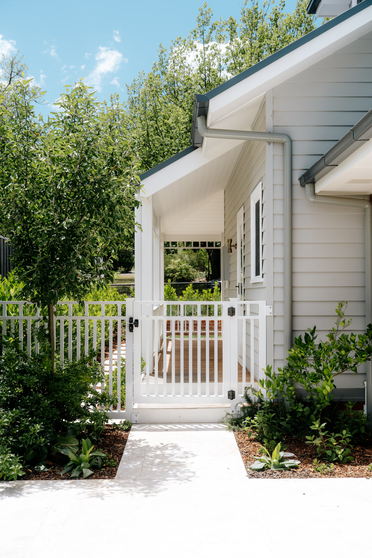 White gated entrance leading to a porch of a modern light gray house surrounded by green plants.