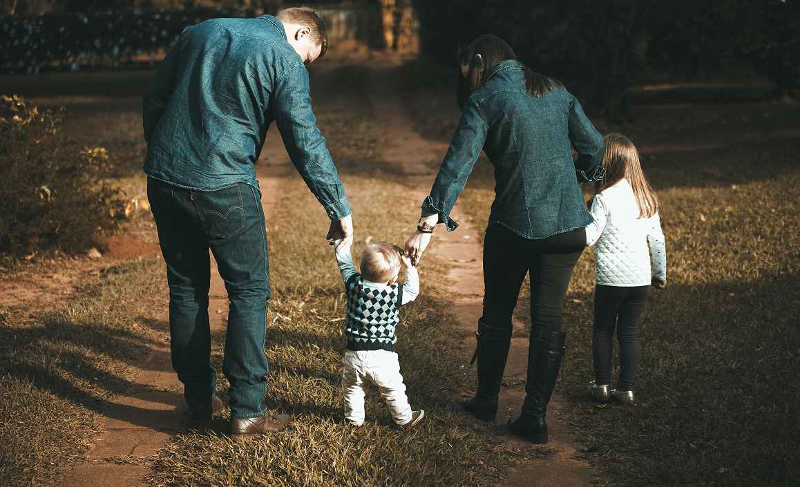 Parents holding hands and helping toddler walk down a dirt path with older child nearby.