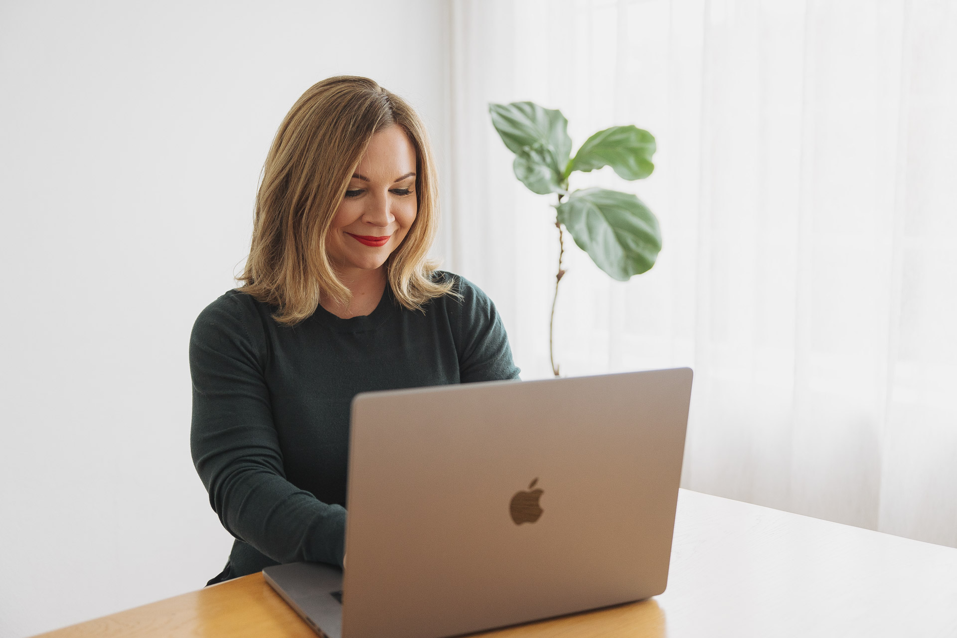 Woman with blonde hair smiling while working on a silver Apple laptop at a wooden table.