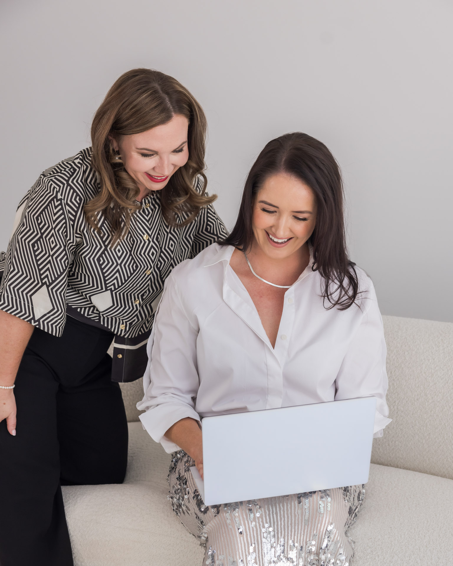 Two women smiling and looking at a laptop while sitting on a light-colored couch.