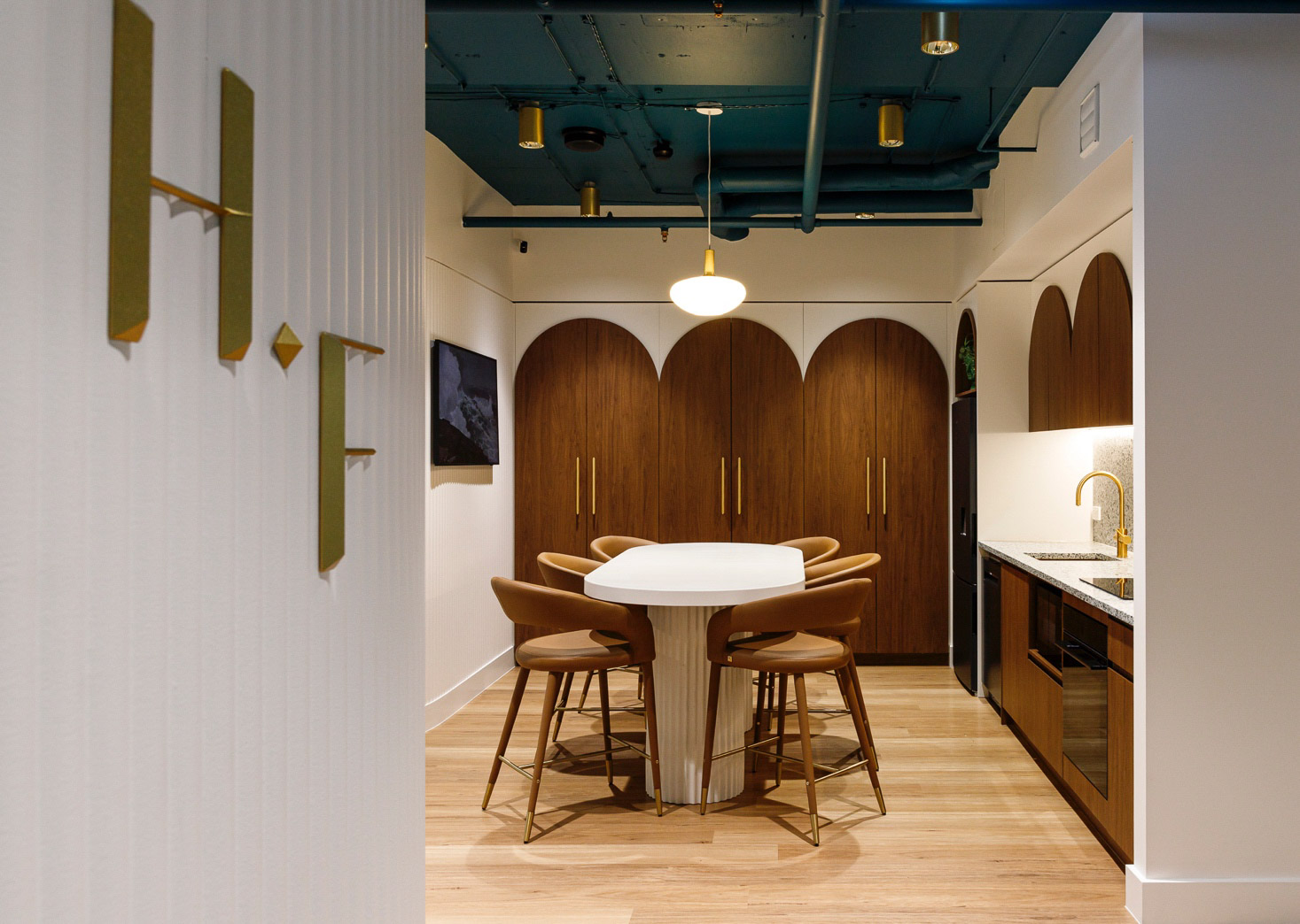 Modern office kitchen with white central table, brown chairs, wooden curved cabinets, and blue ceiling pipes.