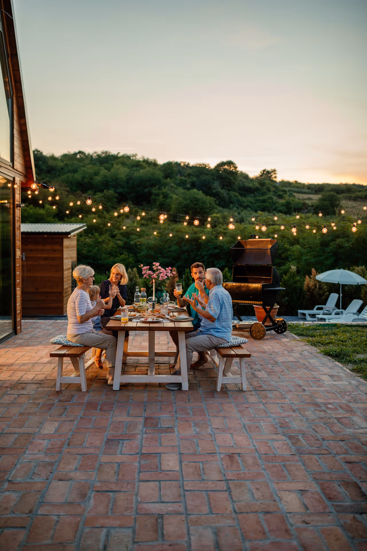 Three generations of family enjoying dinner together at a wooden table outdoors with hanging string lights at sunset.