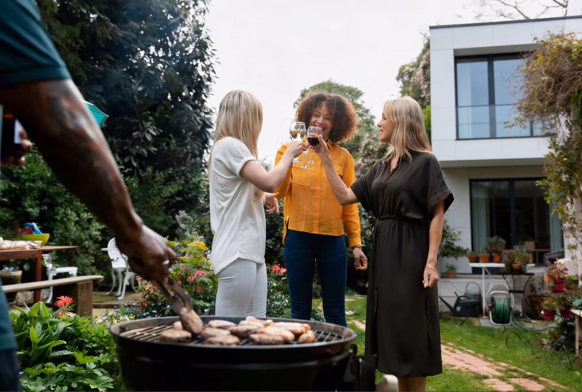 Three women clinking wine glasses outdoors near a barbecue grill with grilled sausages and burgers.
