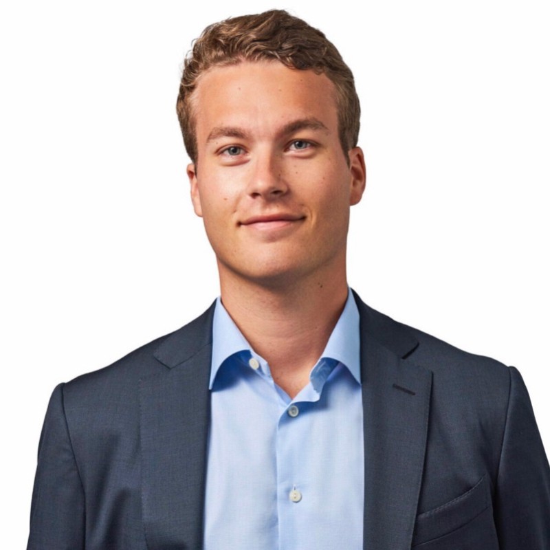 Henry Kamp wearing a dark suit jacket and light blue dress shirt, smiling slightly against a white background.