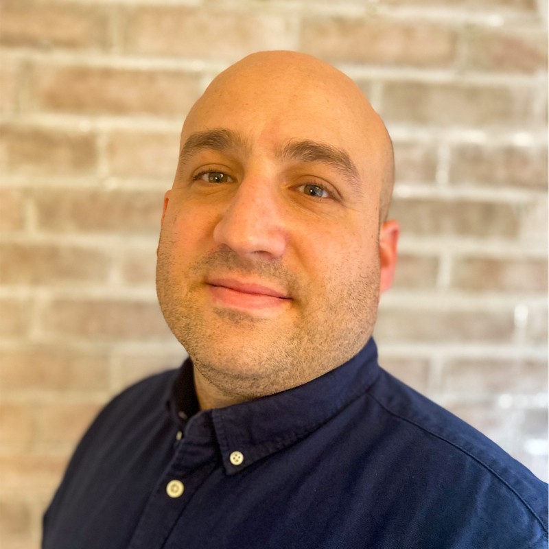 Phil Brandt wearing a navy blue button-up shirt with a blurred brick wall background.