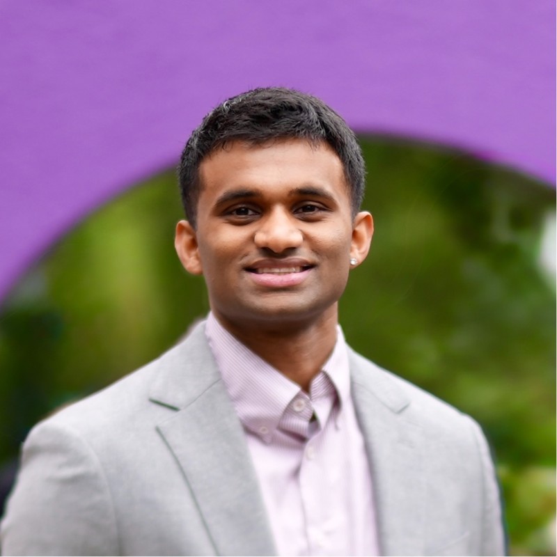 Young man in a light gray blazer and light pink shirt smiling in front of a purple and green blurred background.