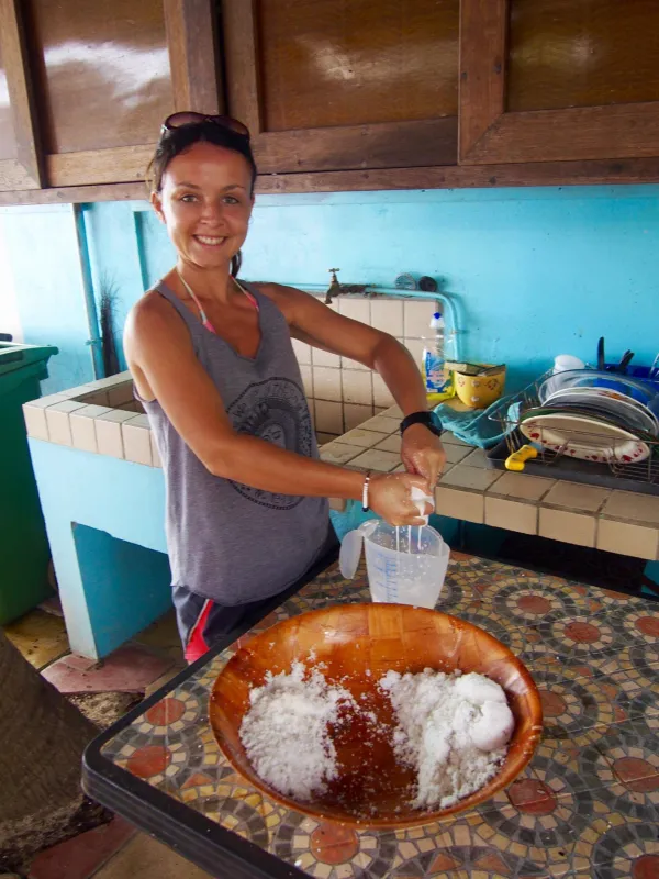 Smiling woman squeezing fresh coconut milk into a measuring jug in a kitchen with a wooden bowl of grated coconut on the tiled table.