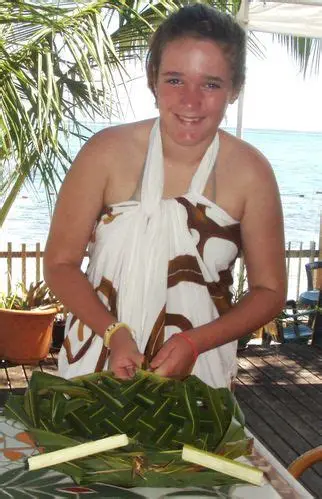 Smiling woman dressed in a white and brown patterned wrap preparing woven green leaves on a table by the beach with palm trees in the background.