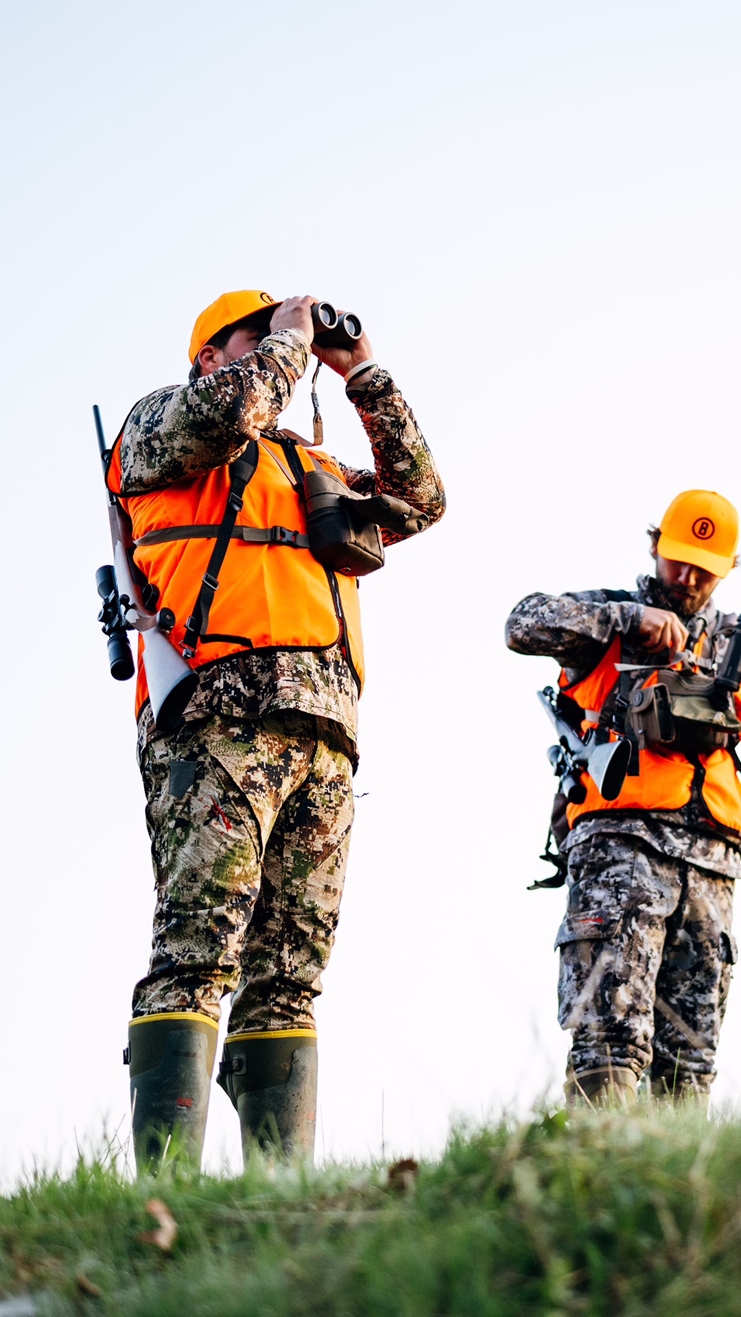 Two hunters in camouflage and orange vests on grassy hill, one looking through binoculars, the other adjusting gear.