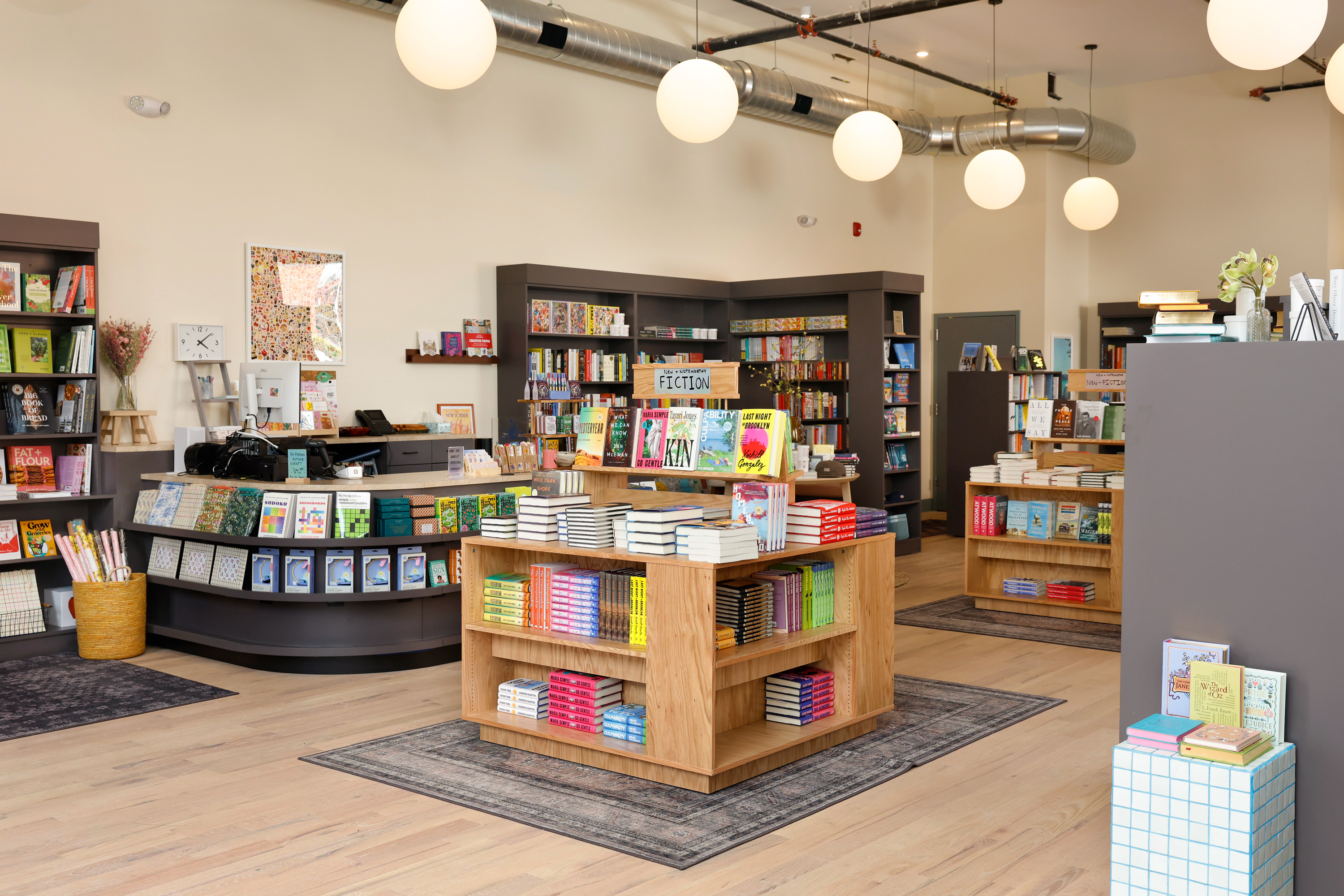 Bright bookstore interior with wooden shelves filled with fiction and non-fiction books and round hanging lights.
