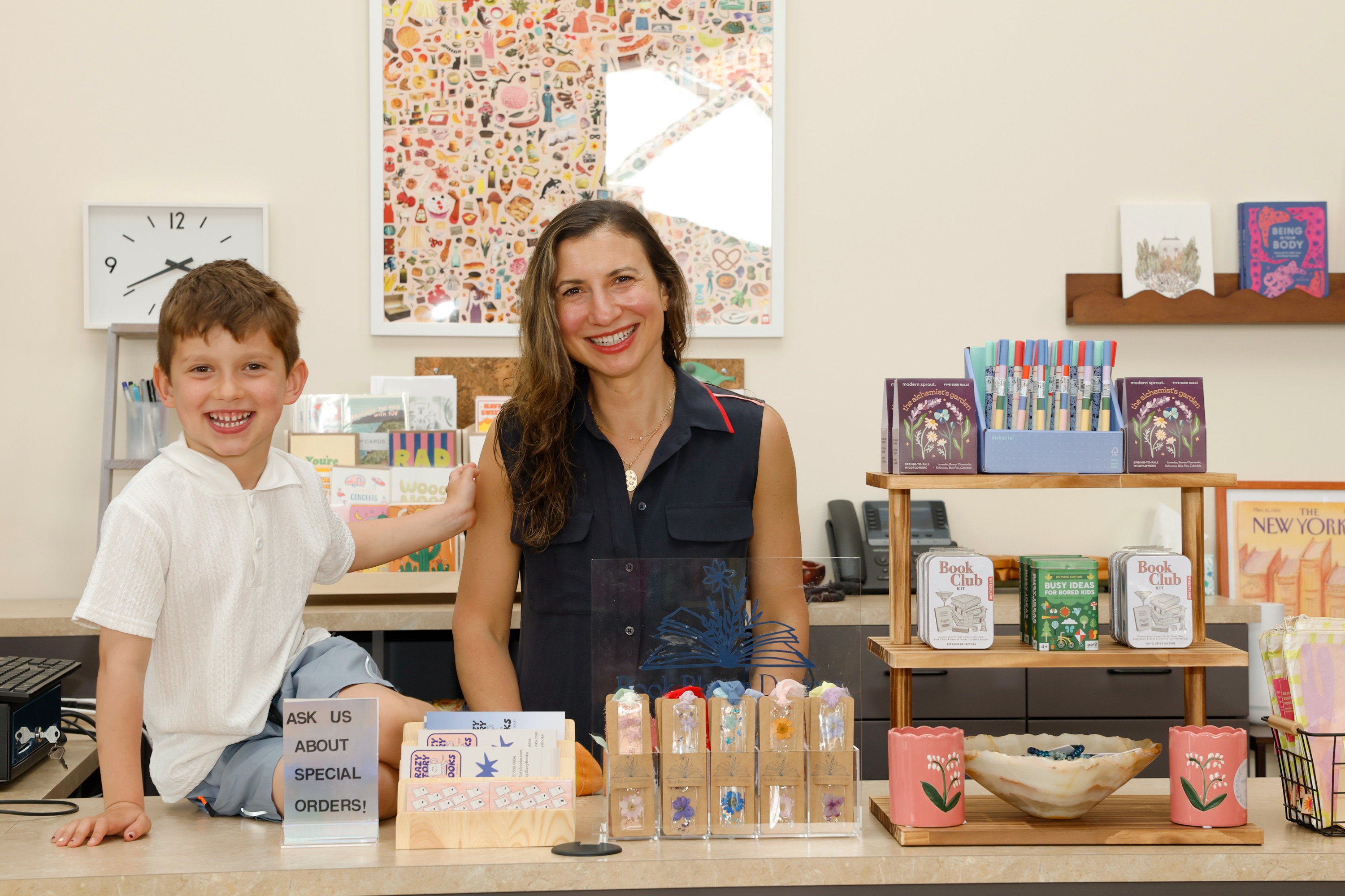 Smiling woman and young boy sitting at a shop counter with colorful stationery and gift items.