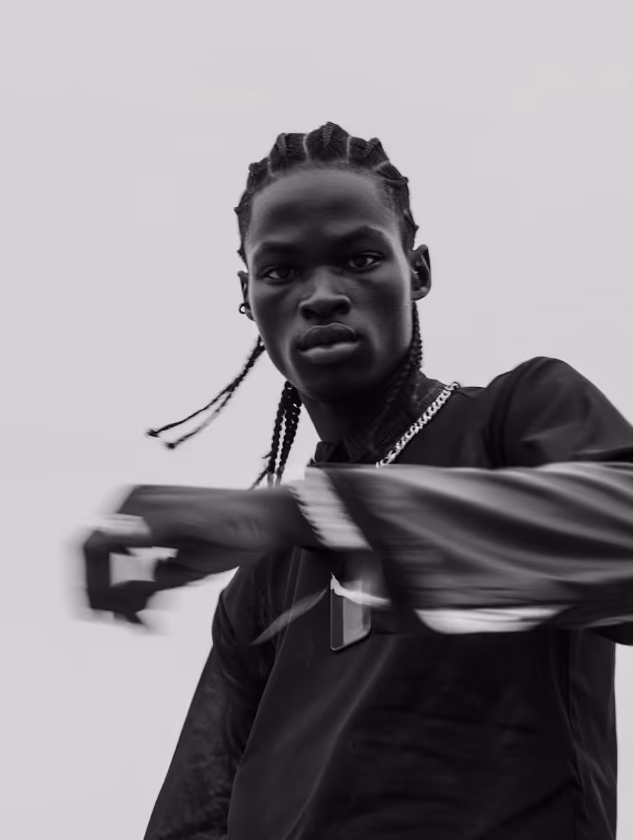 Black and white portrait of a man with braided hair wearing a chain necklace and long-sleeved shirt, reaching out with a blurred hand.