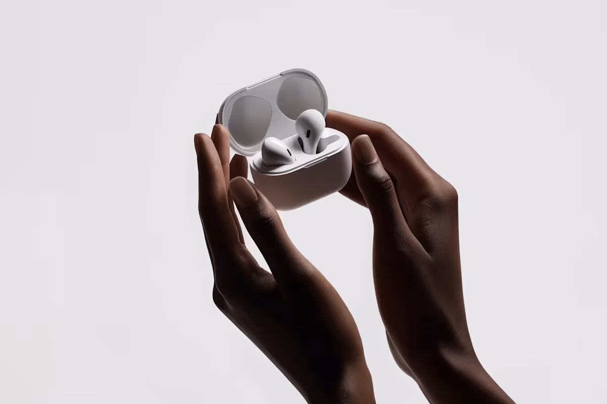 Dark-skinned hands holding an open white case with wireless earbuds inside against a plain light background.
