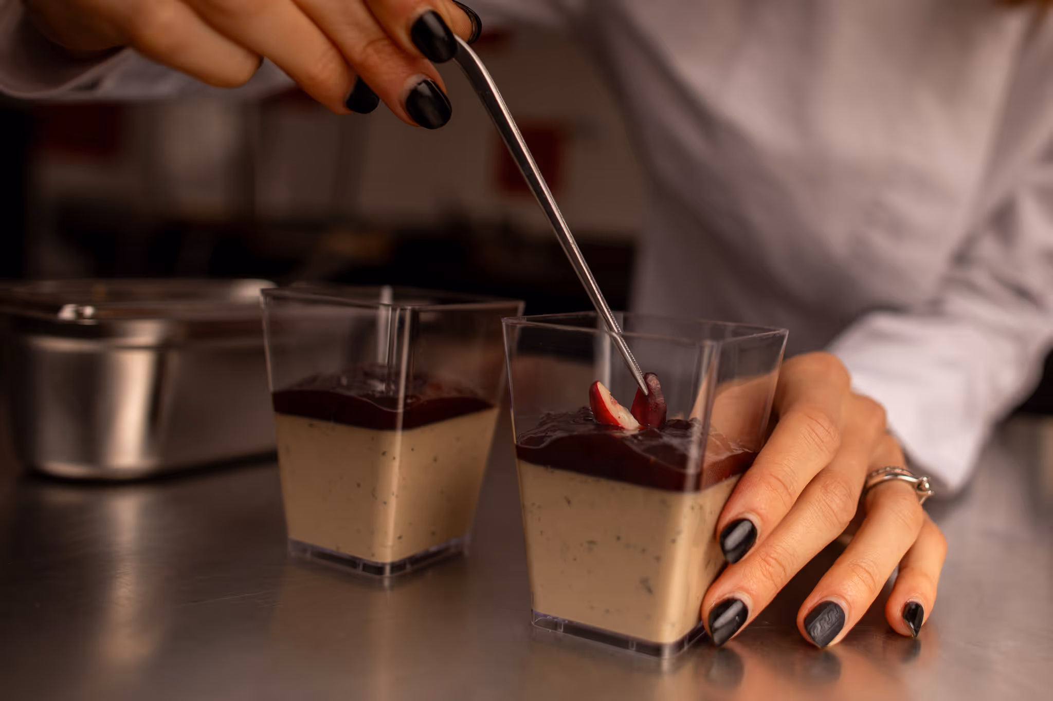 Ruce s černým lakem na nehtech drží a zdobí dezert v průhledné nádobce kovovou lžičkou.