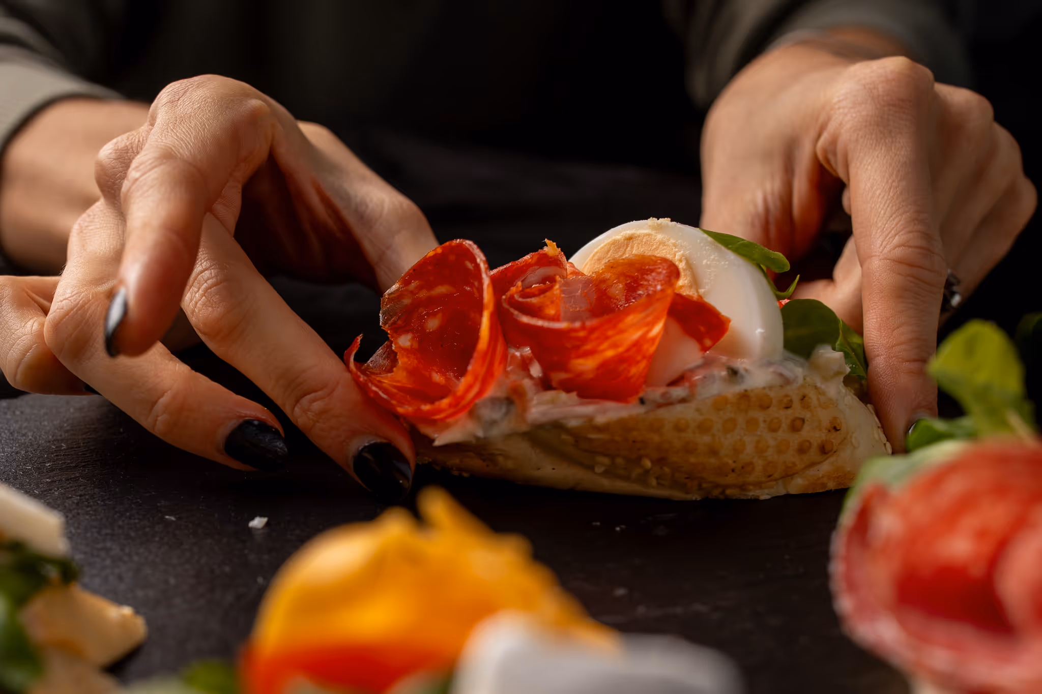 Ruce držící kanapku s plátky salámu, vařeného vejce a zelenými listy na černém povrchu.