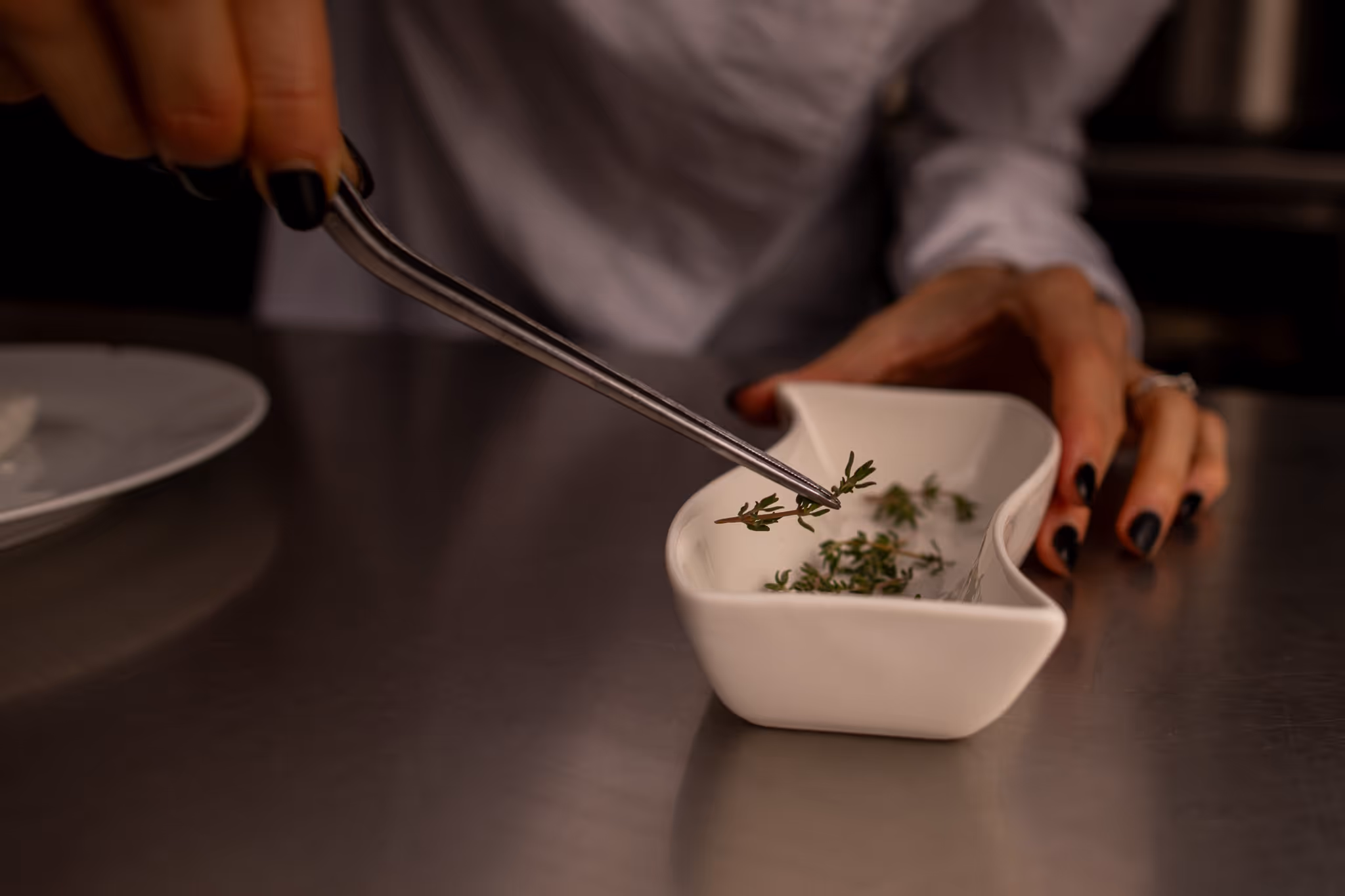 Ruce s černým lakem na nehtech používají pinzetu k pokládání bylinek do malé bílé misky na nerezovém stole.