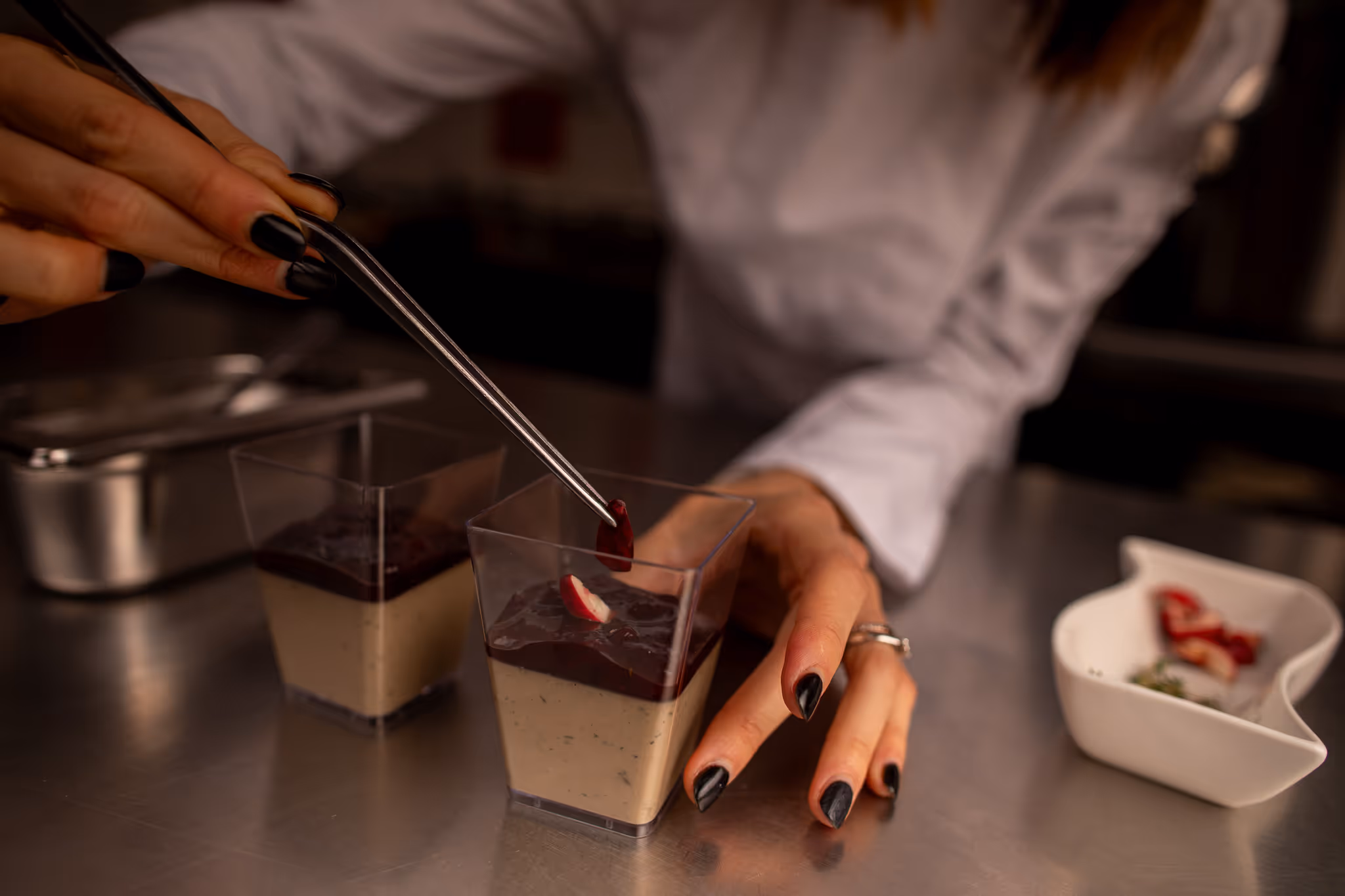 Osoba dekorující dezert krémem a ovocem v průhledných plastových kelímcích na kuchyňském stole.