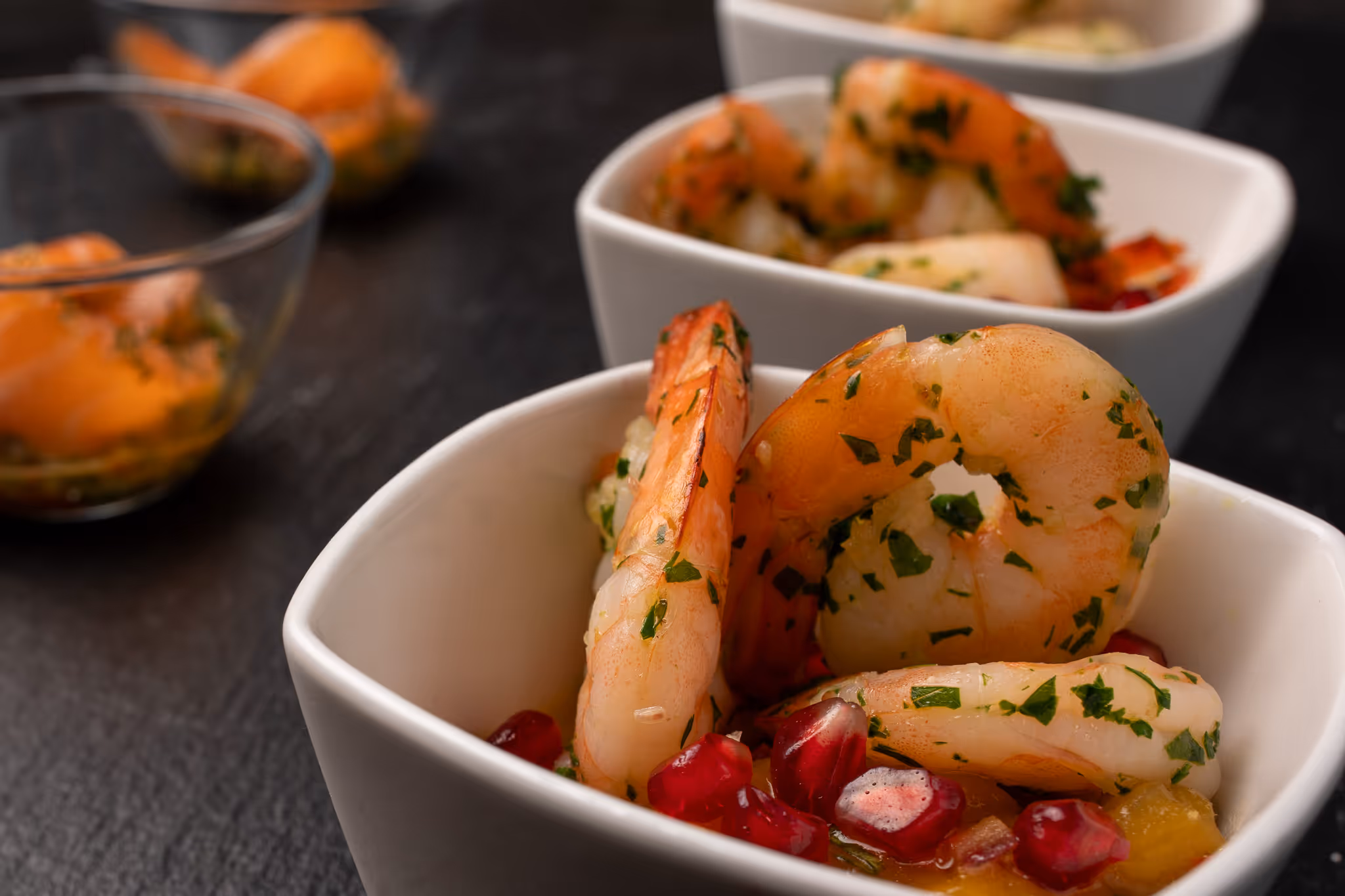 Misky s krevety ochucenými bylinkami a granátovými jablky na tmavém povrchu.