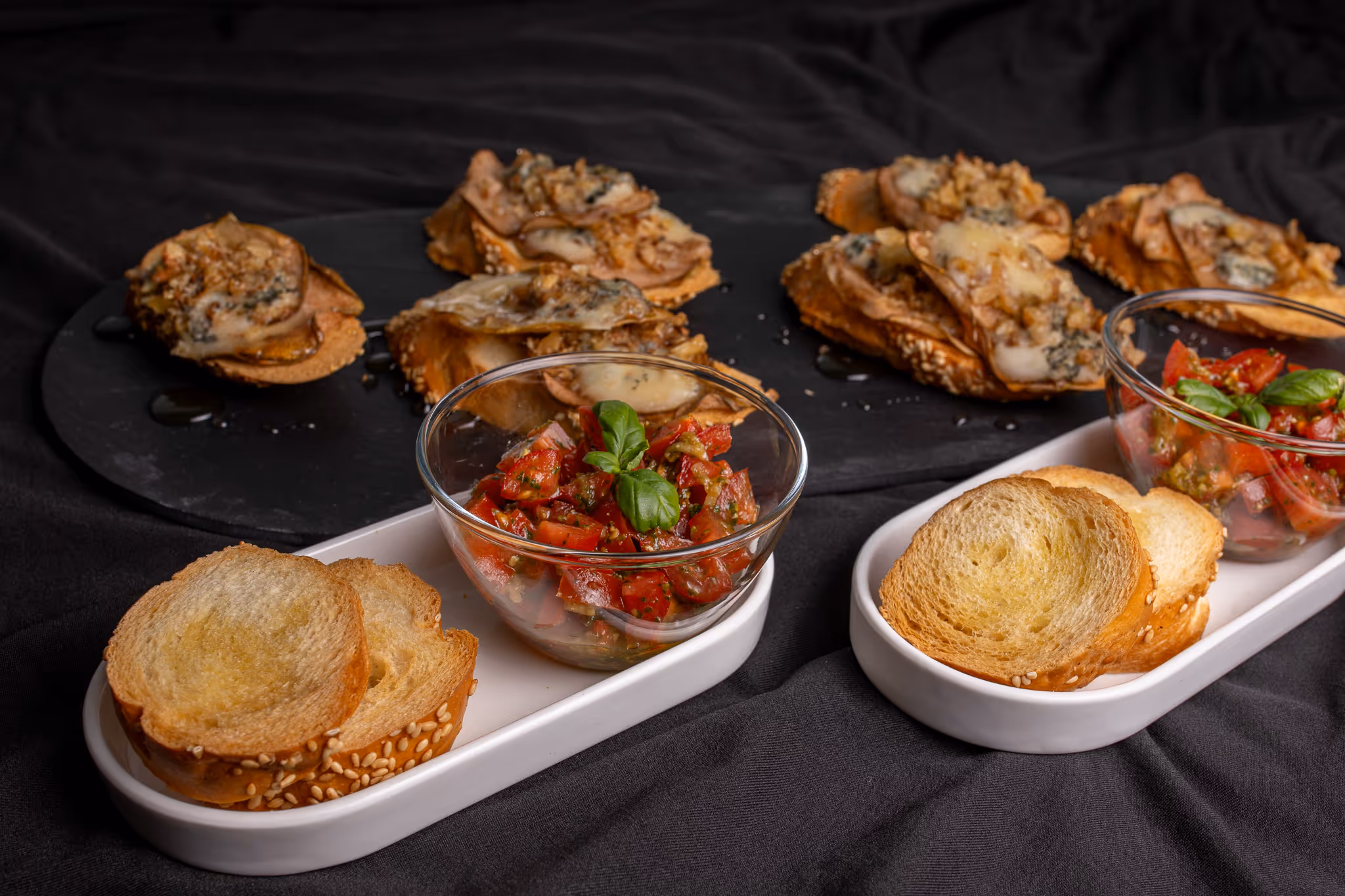 Miska s rajčatovým salátem a bylinkami vedle skladů opečeného chleba a zeleninových křupek s rozplývajícím se sýrem na černém podkladu.