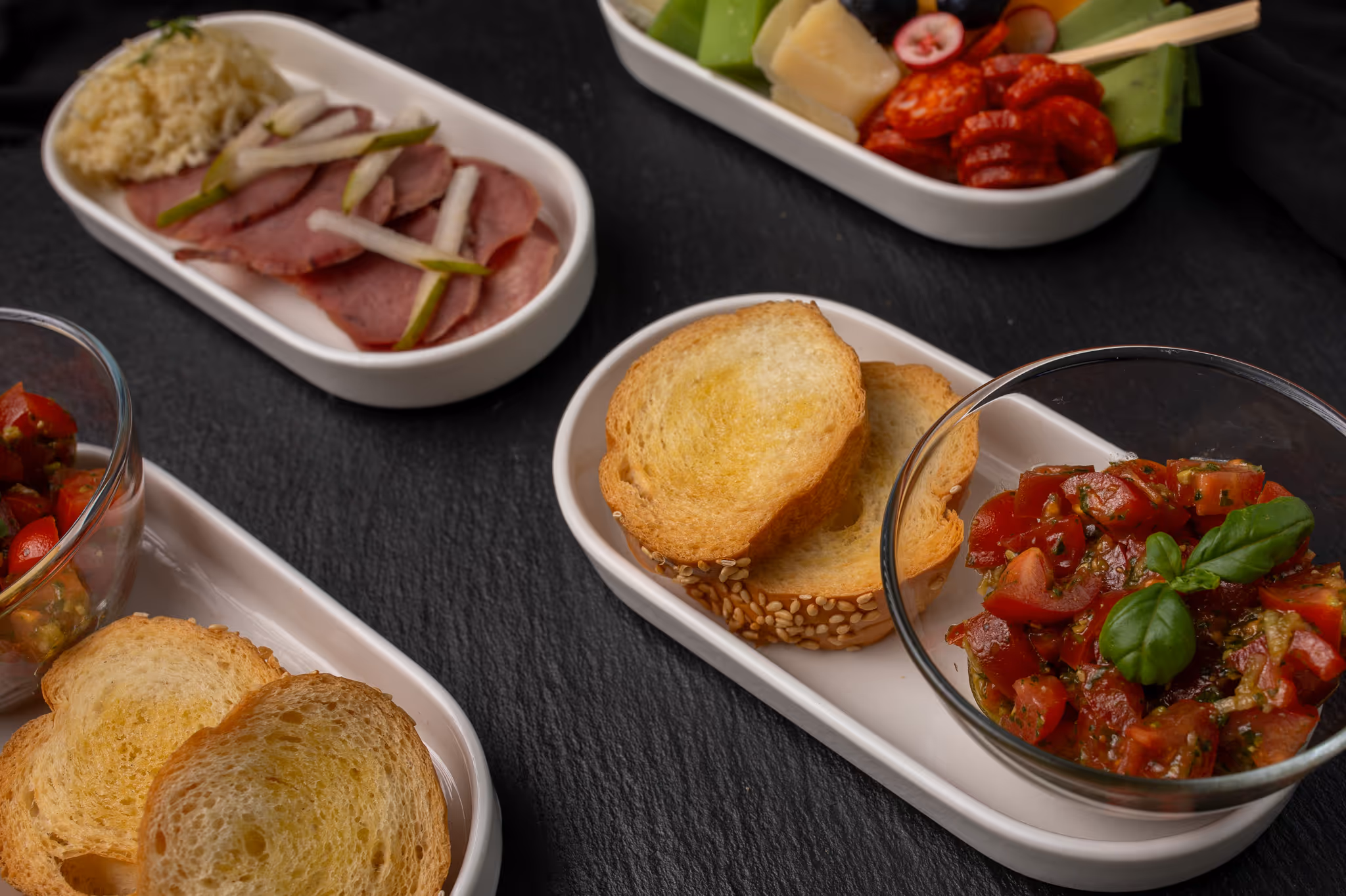 Talířky s plátky pečeného masa, nasekanými rajčaty se zelenými bylinkami a plátky opečeného chleba na černém pozadí.