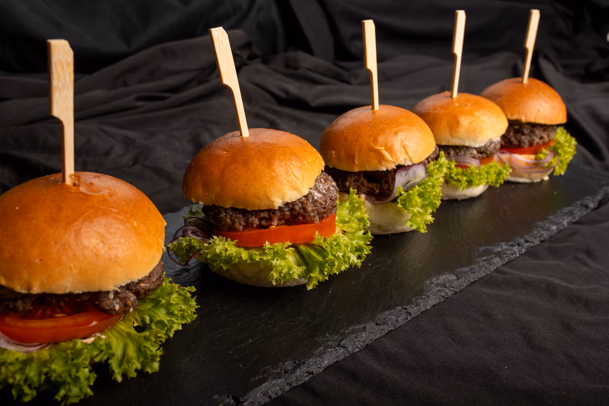 Pět mini hamburgerů s hovězím masem, listovým salátem, rajčetem a cibulí, každý s dřevěnou špejlí, podávané na černém podnose.