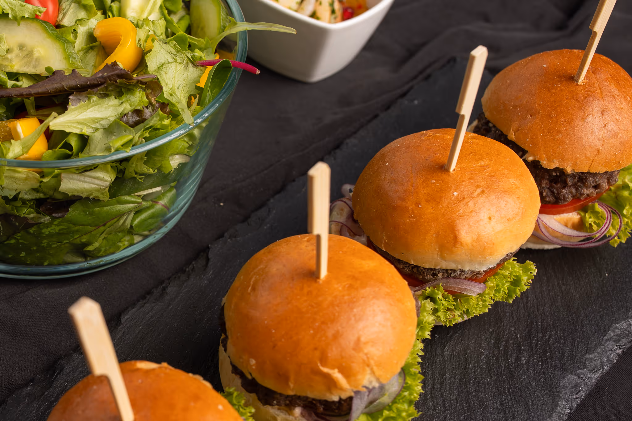 Tři malé hamburgery s hovězím masem, ledovým salátem, cibulí a rajčetem na černé desce vedle misky se salátem.