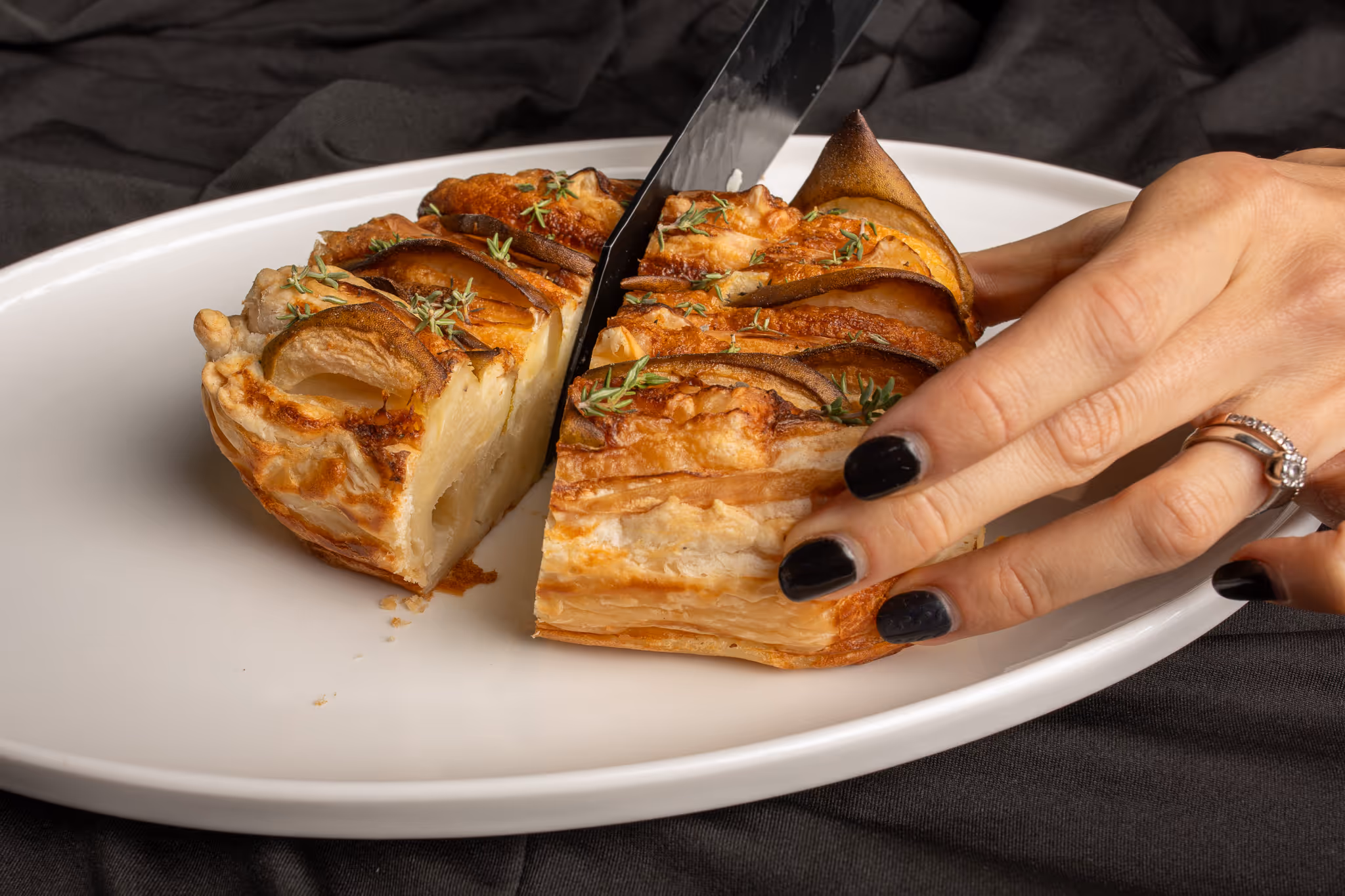 Ruka s černě nalakovanými nehty krájí jablečný koláč ozdobený plátky jablek a tymiánem na bílém talíři.