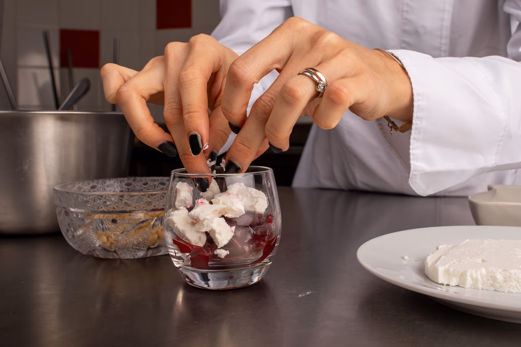 Ruce s černým lakem na nehtech vkládají kousky bílého sýra do skleničky s červenou zeleninou.