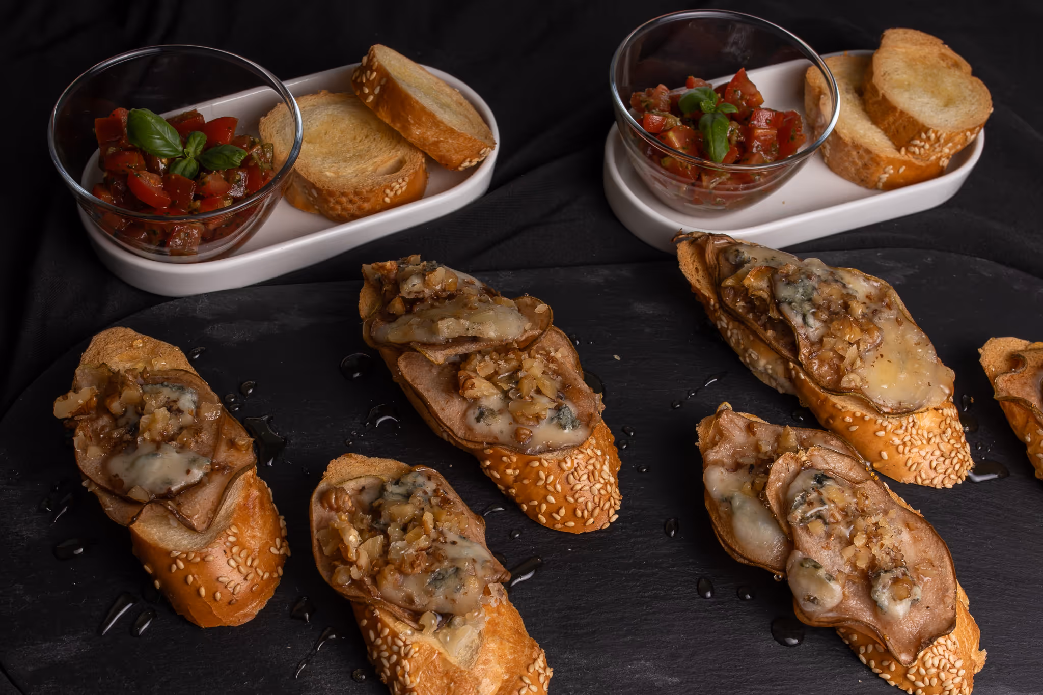 Topinky s plátky hrušek, rozpuštěným sýrem a nasekanými ořechy na černé podložce, vedle misek s rajčatovou salsou a plátky opečeného chleba.