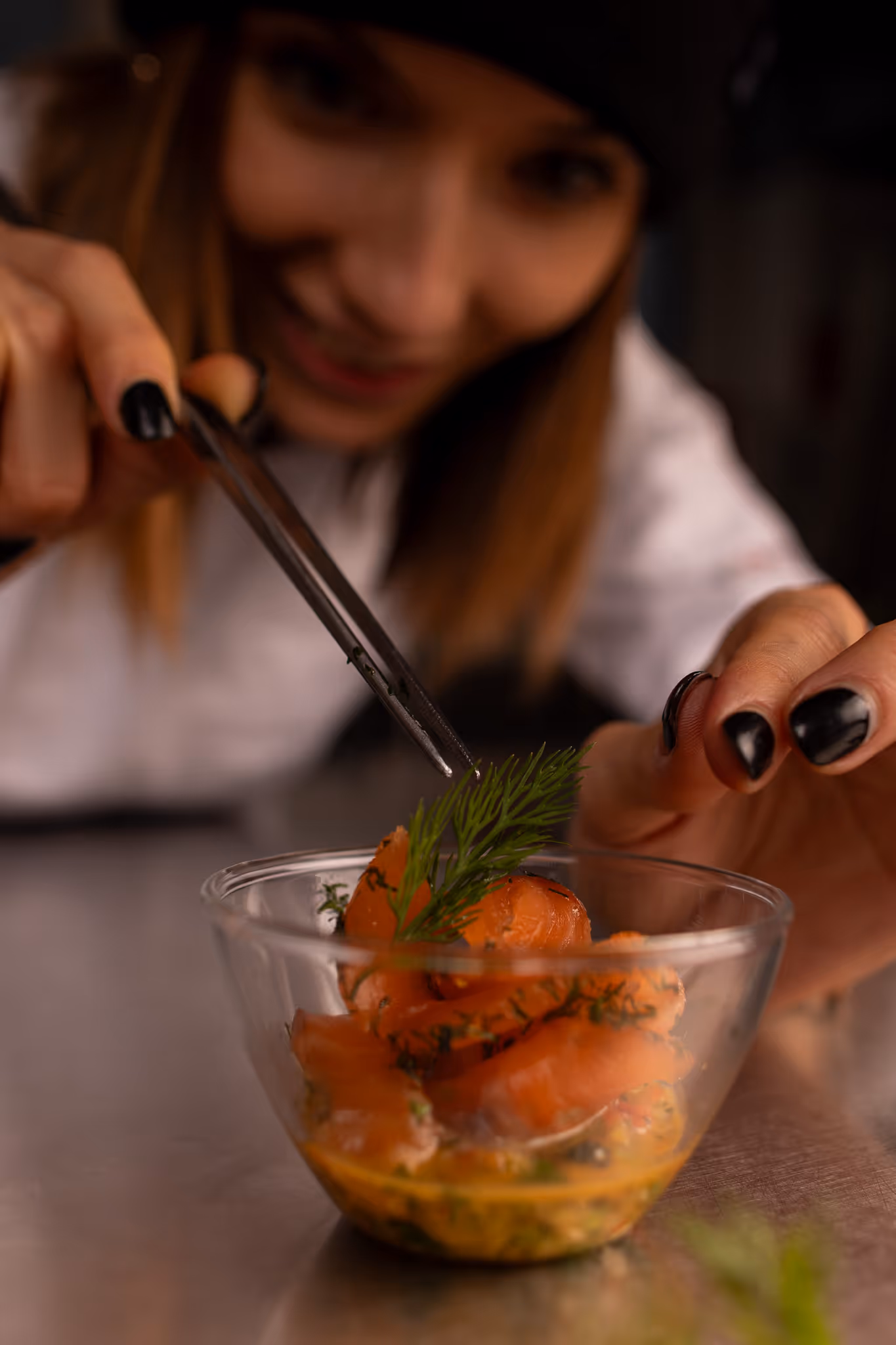 Žena s pinzetou zdobí pokrm plátky lososa a koprem v průhledné misce.
