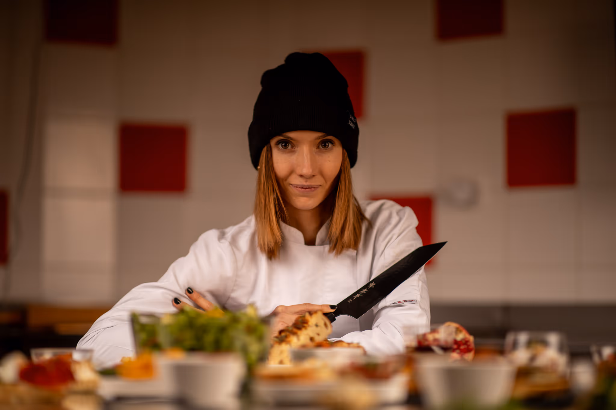 Žena kuchařka v bílé šéfské zástěře a černé čepici drží velký nůž v kuchyni před talíři s jídlem.