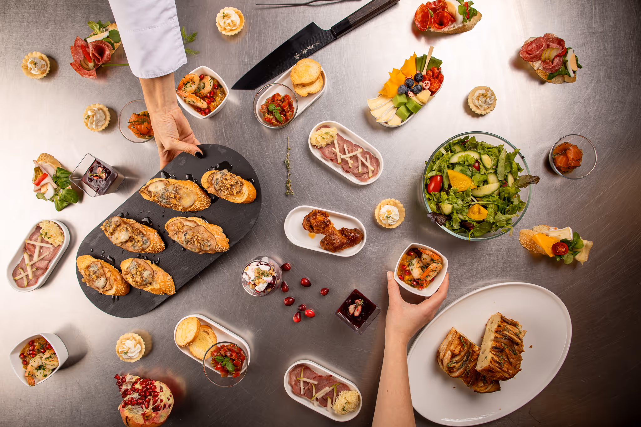 Různé lahodné předkrmy, salát a dezerty pečlivě naaranžované na kovovém stole s rukama držícími talíře s jídlem.